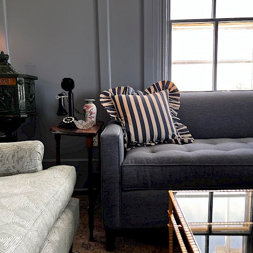 A cozy living room with a grey sofa, striped pillow, side table with a lamp and mug, and a glass coffee table in front of a window.