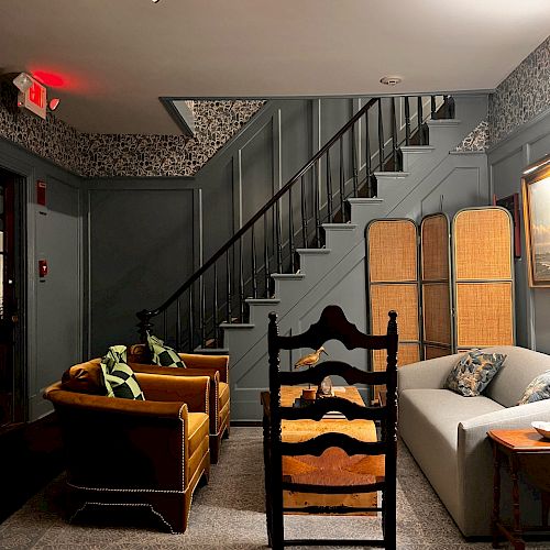 A cozy living area with a staircase, yellow sofa, and chairs around a table. A divider and a grey sofa are also present under warm lighting.