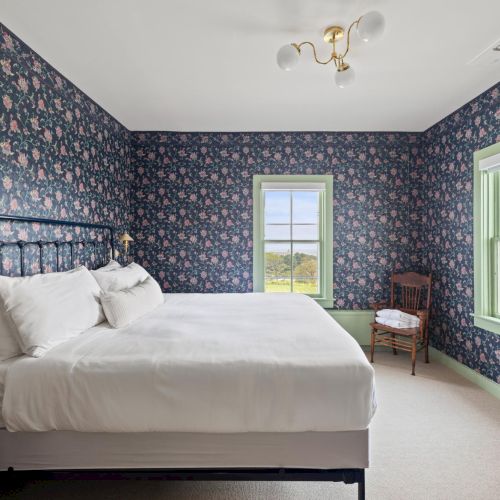 A bright bedroom with a large bed, light gray headboard, two blue accent walls, two windows with sheer curtains, and a ceiling fan.