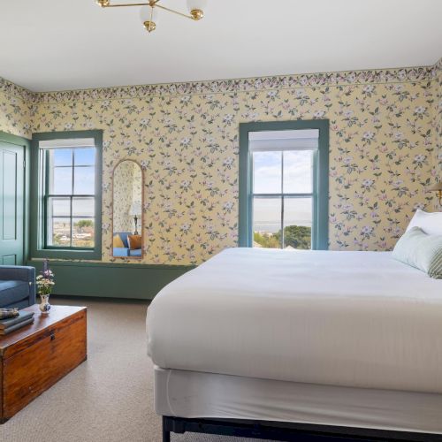 A bright hotel room with two beds, beige patterned wallpaper, a wooden dresser, a sofa, a window view, and soft natural light.