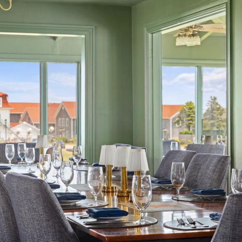 A cozy dining area with a long table set for a meal, hanging lights, and large windows letting in natural light.