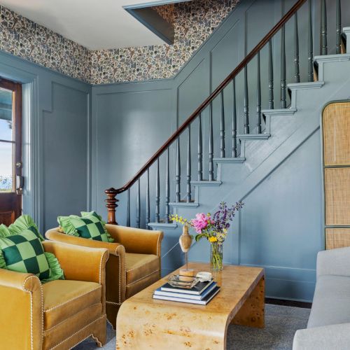 A bright, cozy living room with yellow sofa, teal armchairs, a wooden staircase, and warm natural light. Top it at 140 characters, always ending the sentence.