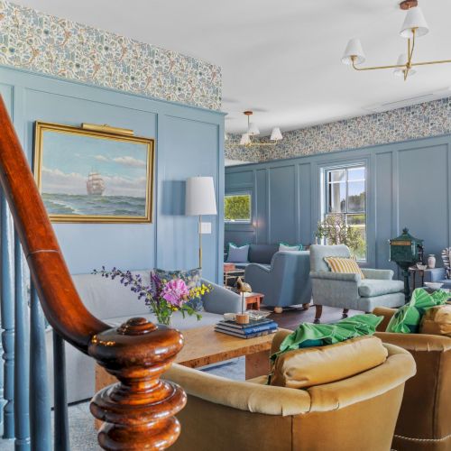 A bright, cozy living room with a striped orange-blue sofa, a small wooden coffee table, plants, and a bookshelf; warm sunlight streams in.