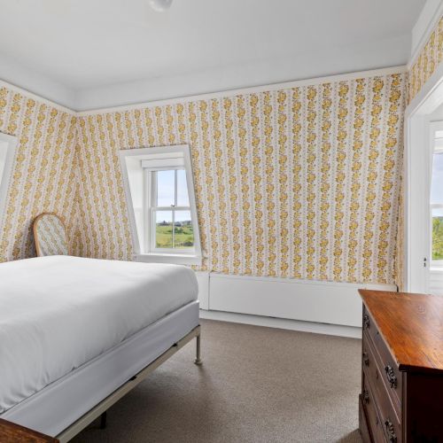 A bright, cozy bedroom with a large window, soft gray bed, mustard accent wall, and warm wooden floors.