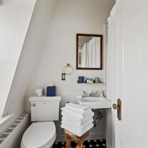 A compact bathroom with a white toilet, pedestal sink, towel stack, mirror cabinet, small shelf, blue accents, and a window letting in light, neat.