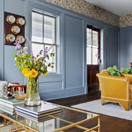 A stylish living room with blue paneled walls, a beige sofa, mustard chairs, glass coffee table, and fresh flowers in a vase.