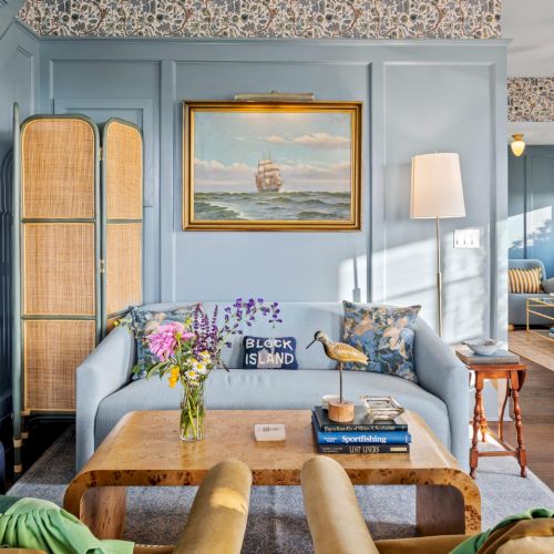 Colorful coastal living room with blue walls, wicker screens, a wooden coffee table, sailboat art, bright flowers, and mismatched, comfy seating. End.