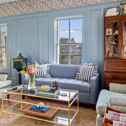 A bright living room with blue walls, large windows, a blue sofa, striped cushions, a glass coffee table, and a tall wooden cabinet, stylish and cozy.